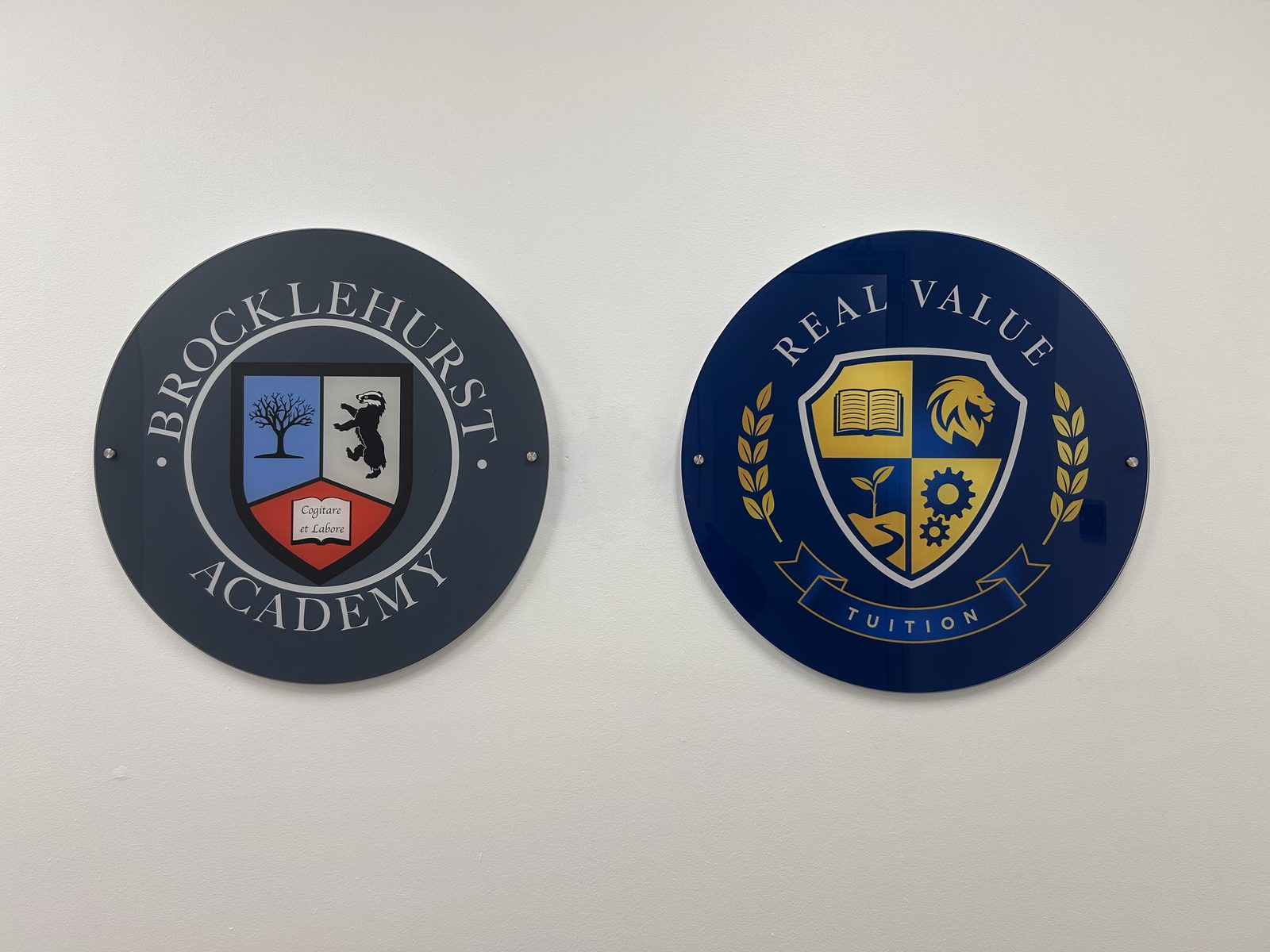 Two round signs side by side on a white wall at the shared Burwood centre: the Brocklehurst Academy crest (motto ‘Cogitare et Labore’) on the left and the Real Value Tuition crest on the right